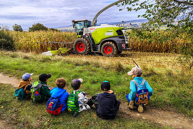 Kinder sitzen am Rand eines Feldes und beobachten einen Maishäcksler bei der Arbeit.