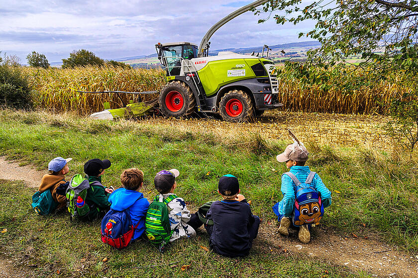 Kinder sitzen am Rand eines Feldes und beobachten einen Maishäcksler bei der Arbeit.