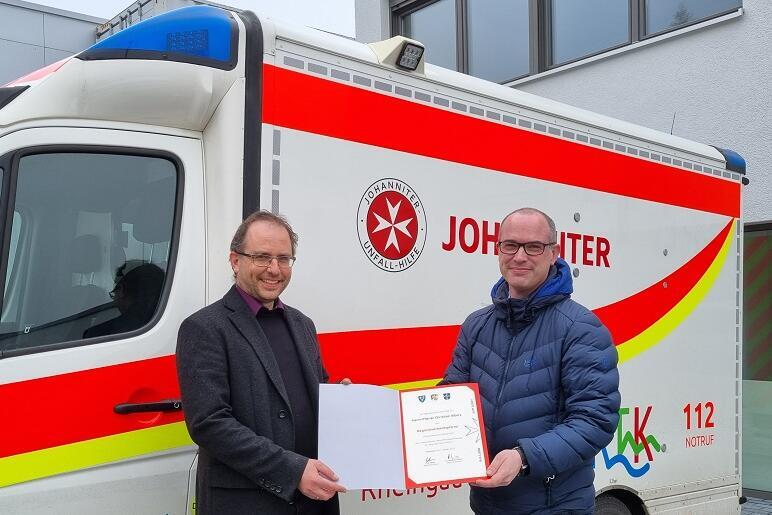 Ulf Weyer überreicht dem neuen Johanniter-Pfarrer Christian Albers seine Ernennungsurkunde vor einem Rettungswagen.