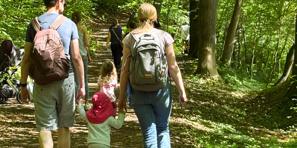 Ein Mann und eine Frau gehen mit einem Kind in der Mitte und an der Hand haltend auf einem Waldweg.