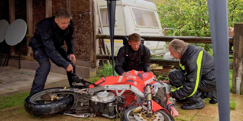 Drei Motorradfahrer kümmern sich um einen verunfallten Motorradfahrer, der halb unter seinem Motorrad liegt. Einer scheint gerade das Motorrad aufstellen zu wollen, einer legt die Hand auf den Arm des Verunfallten, der dritte greift an den Helm.