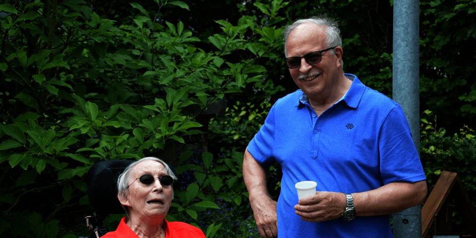 Eine Frau in einem roten Oberteil sitzt in einem Rollstuhl im Garten. Neben ihr steht ein Mann in einem blauen Hemd.
