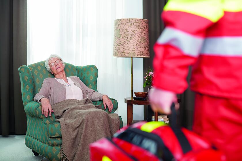 Eine ältere Dame liegt ohnmächtig in einem Sessel, sie trägt einen Hausnotruf Knopf um den Hals. Vor ihr steht eine Rettungskraft in Uniform.
