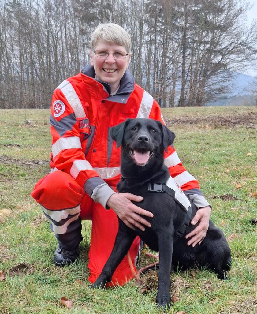 Eine Johanniter-Rettungshundeführerin kniet in roter Einsatzkleidung auf einer Wiese und hält ihren schwarzen Rettungshund im Arm. Beide lächeln in die Kamera, im Hintergrund sind Bäume und ein leicht bewölkter Himmel zu sehen.