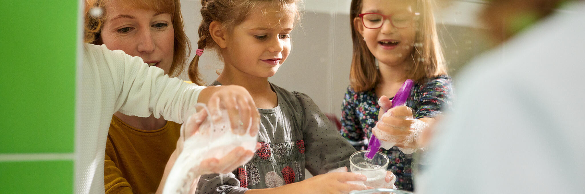 Eine Gruppe Mädchen spielt gemeinsam mit einer Erzieherin mit Wasser und Schaum am Waschbecken.