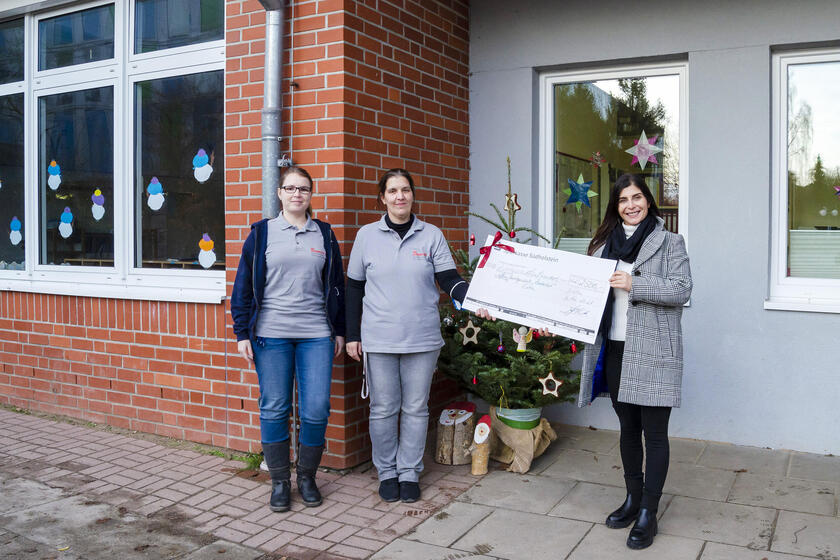 Irina Lüth und Claudia Lejeune (Johanniter-Unfall-Hilfe) nehmen den Spendencheck von Güler Kirk (Geschäftsführerin Gaumenschmaus) vor der Offenen Ganztagsschule Zauberhut entgegen.