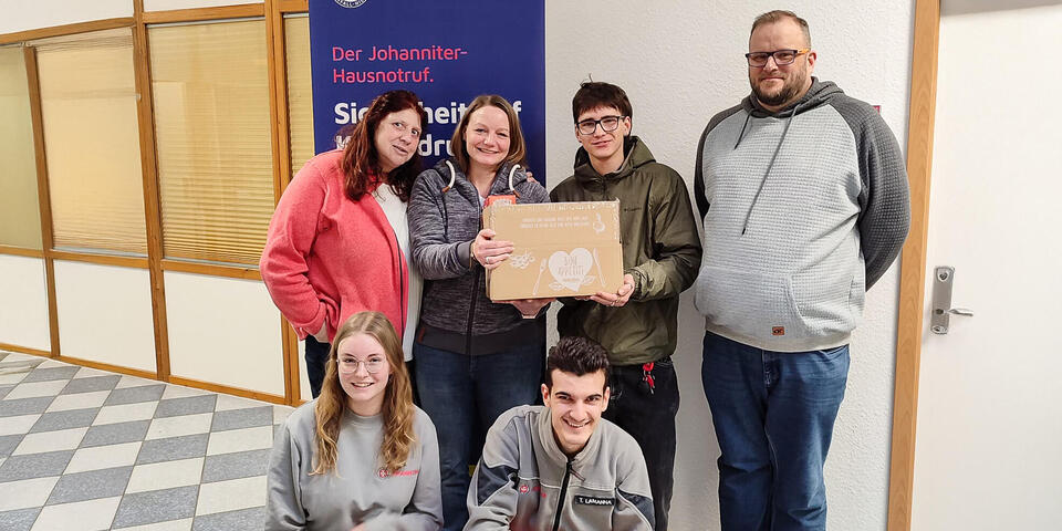 Zwei Frauen, ein junger Mann mit einem Päckchen, ein weiterer Mann vor einem Hausnotruf-Roll-Up. Davor knien eine junge Frau und ein junger Mann in Einsatzkleidung.