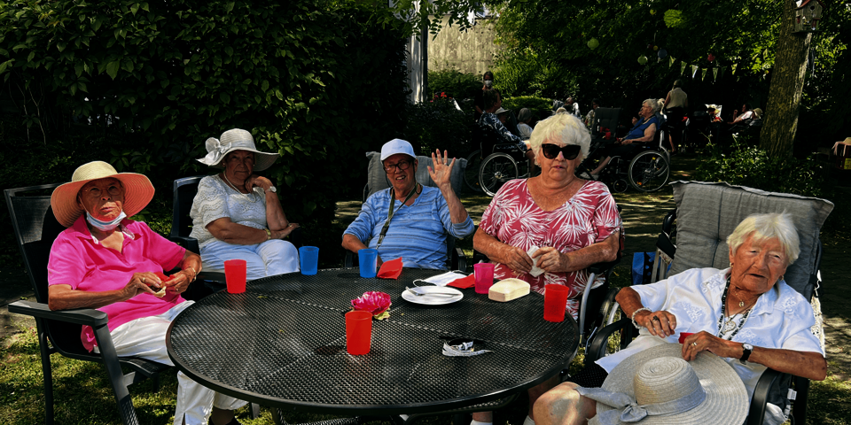 fünf Seniorinnen sitzen mit Sonnenhüten oder Sonnenbrillen an einem Gartentisch.