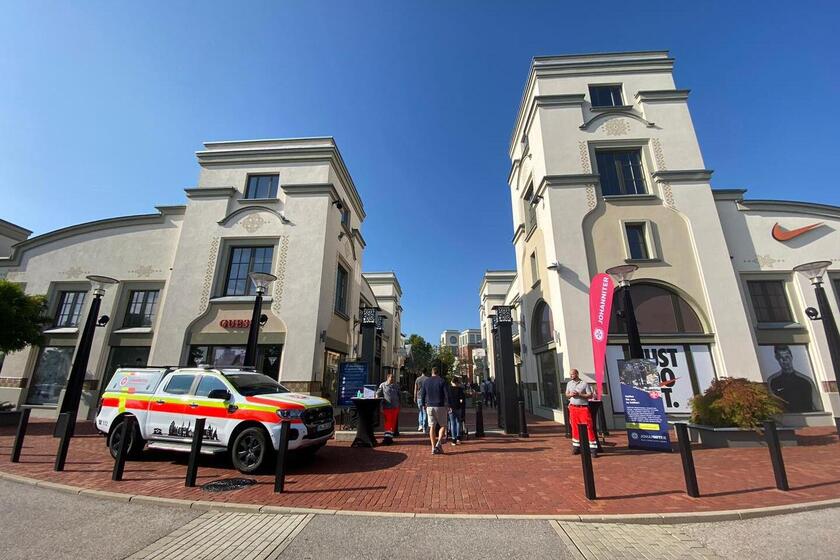 Johanniter-Einsatzfahrzeug und Infostand vor einem Einkaufszentrum bei sonnigem Wetter.