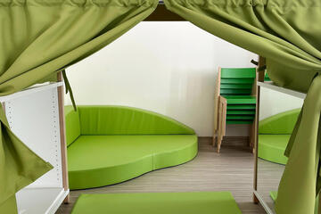 eine Kuschelecke, wie ein kleines Häuschen mit Vorhängen an den Seiten, einer Matte auf dem Boden und links Regale für Spielzeug, rechts ein Spiegel, alles in Grün gehalten