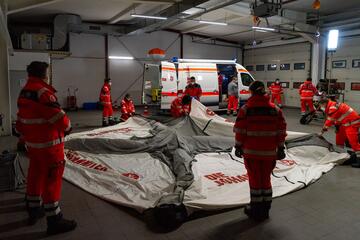 Mehrere Johanniter-Einsatzkräfte bauen gemeinsam in einer Halle ein großes Zelt mit der Aufschrift „Die Johanniter“ auf. Im Hintergrund steht ein Rettungswagen.