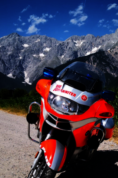 Zwei Motorräder der Johanniter mit roter und silberner Lackierung stehen auf einer Landstraße vor einer beeindruckenden Bergkulisse unter blauem Himmel.