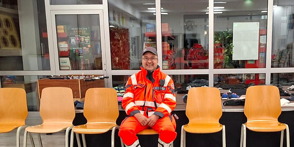 Ein Mann in Einsatzkleidung sitzt auf einem Stuhl in einer Stuhlreihe. Er befindet sich in einer Aula.