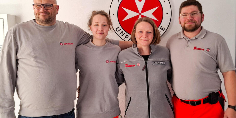 Zwei Frauen und zwei Männer in Johanniter-Oberteilen in grau. sie stehen vor einer Wand mit einem Johanniter-Logo