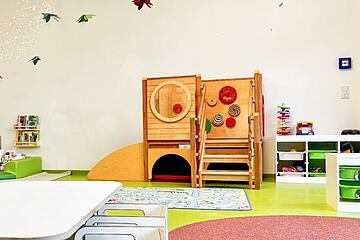 Ein heller Spielraum mit einer kleinen Holzkletterstation, bunten Teppichen und niedrigen Regalen mit Spielmaterial.