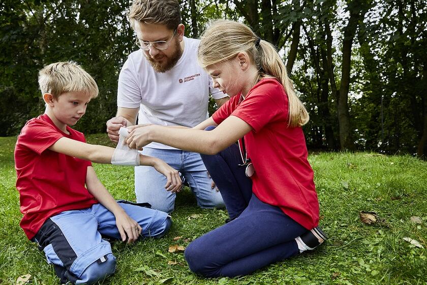 Zwei Kinder üben das Anlegen eines Verbands mit einem Jugendgruppenleiter.