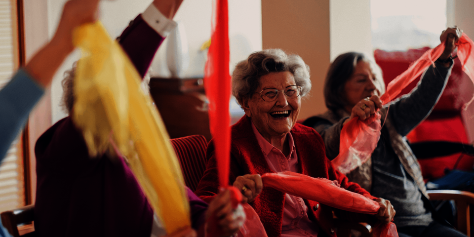  Mehrere Seniorinnen sitzen lachend und bewegen bunte Tücher im Gemeinschaftsraum.
