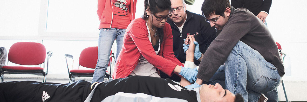 Teilnehmende eines Erste-Hilfe-Kurses üben die Anwendung von Erste-Hilfe-Maßnahmen an einem liegenden Teilnehmer. Ein Ausbilder unterstützt sie dabei.