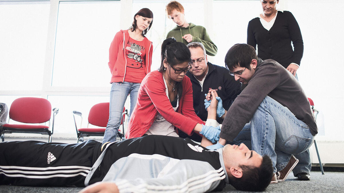 Teilnehmende eines Erste-Hilfe-Kurses üben die Anwendung von Erste-Hilfe-Maßnahmen an einem liegenden Teilnehmer. Ein Ausbilder unterstützt sie dabei.