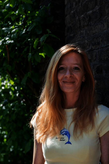 Porträt einer lächelnden Frau vor einer Mauer mit Grün im Hintergrund.