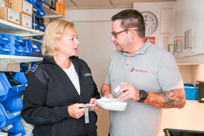 Eine Frau und ein Mann stehen in einem Lager. Sie hält ein Hausnotruf Armband in der Hand und er ein Hausnotrufgerät.