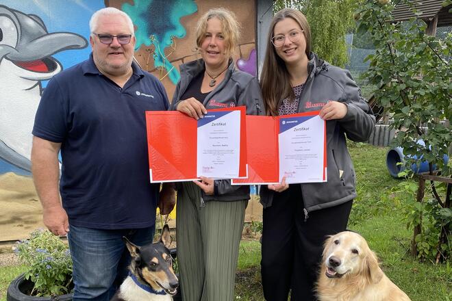 Drei Erwachsene stehen mit zwei Hunden vor einer bunt bemalten Wand im Garten der Kita Hoppetosse in Schwabach. Zwei der Erwachsenen halten Zertifikate in den Händen. Die beiden Hunde, ein Collie-Mischling und ein Golden Retriever, sitzen entspannt auf dem Rasen.