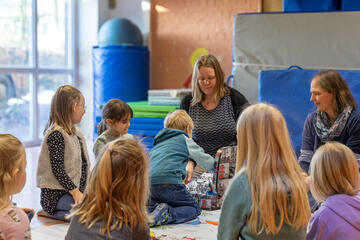 Die Kinder sitzen im Kreis um die Erzieherinnen. In der Mitte steht ein Koffer mit verschiedenen Utensilien, die für den Unterricht gebraucht werden. Ein Kind greift in den Koffer.