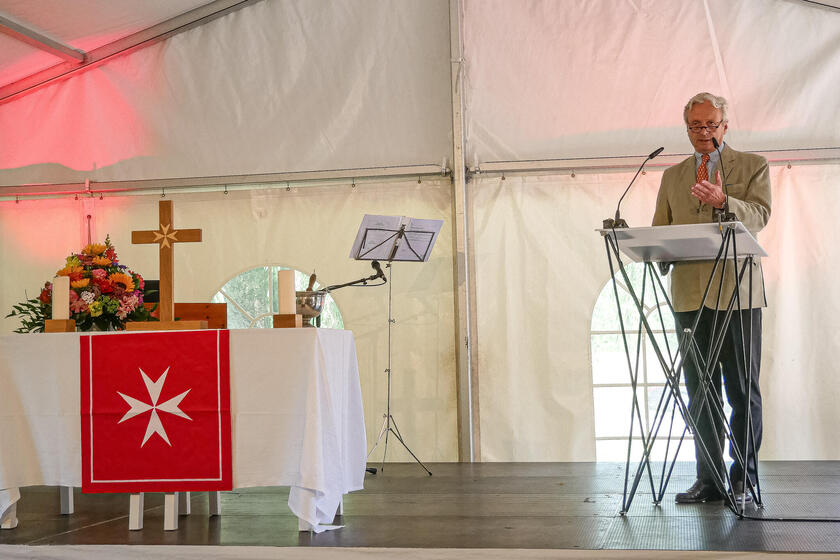 Ein Mann spricht am Rednerpult neben einem Altar mit Blumen, Kreuz und rotem Tuch mit Johanniterkreuz.