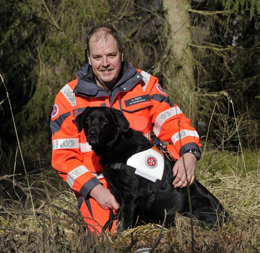 Ein Johanniter-Rettungshundeführer sitzt in leuchtender Einsatzkleidung im Wald und hält seinen schwarzen Rettungshund mit weißer Einsatzweste nah bei sich. Beide blicken aufmerksam in die Kamera. Im Hintergrund sind Bäume und Sträucher zu sehen.