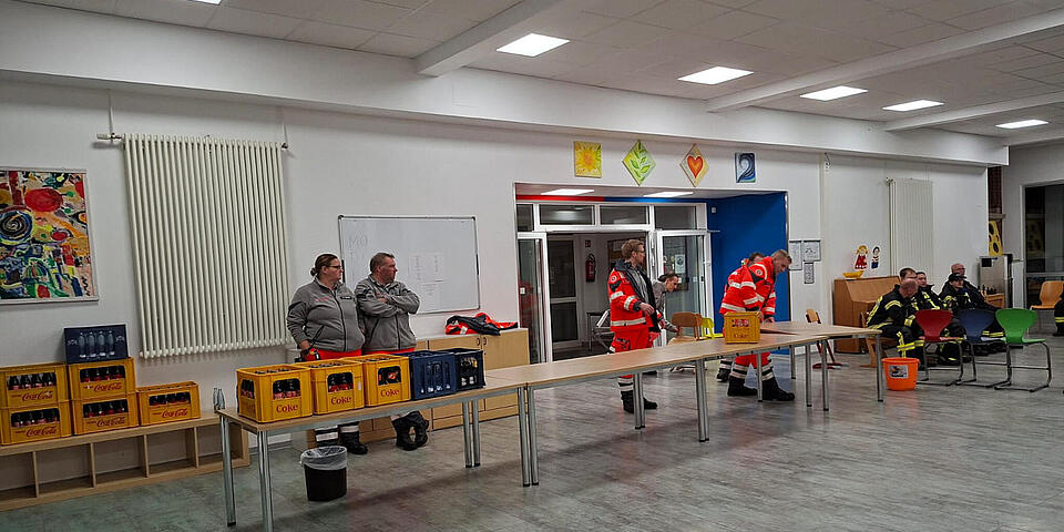 Fünf Menschen in Johanniter-Einsatzkleidung an Tischen mit Getränken in einer Aula.