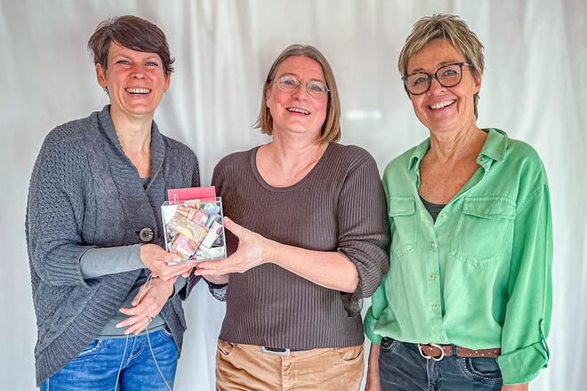 Drei Frauen stehen lächelnd vor einem weißen Vorhang und halten eine kleine Spendenbox in der Hand.