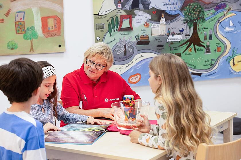 Johanniter-Ehrenamtliche sitzt mit drei Kindern an einem Tisch und begleitet ein Trauergespräch im Rahmen von „Lacrima“. Im Hintergrund hängt ein bunt gemaltes Bild mit Symbolen zu Trauer, Trost und Erinnerung.