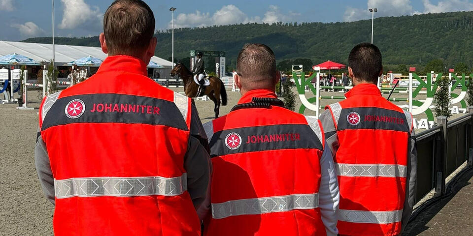Drei Männer mit Johanniter-Einsatzkleidung stehen an einem Reitplatz und schauen einem Reiter zu.