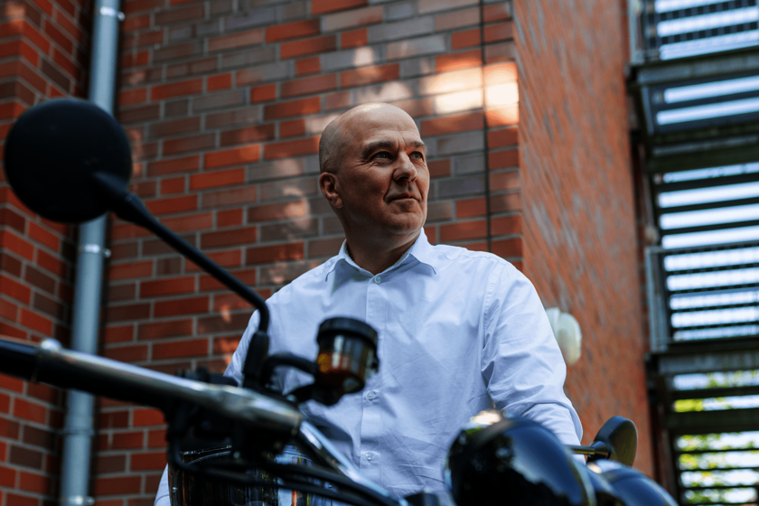 Ein Mann im weißen Hemd steht neben einem Motorrad und hält einen Helm