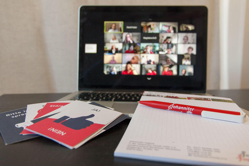 Ein Laptop mit einer Videkonferenz. Im Vordergrund befinden sich Stimmkarten und ein Block sowie ein Johanniter-Jugend-Kugelschreiber.