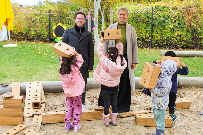 Ein Mann und eine Frau stehen in einem großen Sandkasten. Vor ihnen halten Kinder große Bausteine in die Höhe.