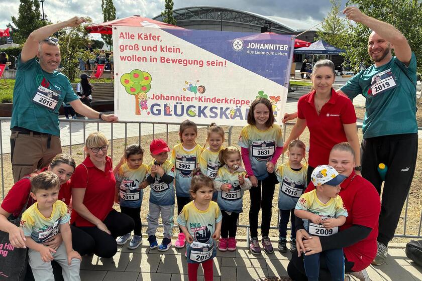 Kinder der Johanniter-Kinderkrippe „Glückskäfer“ platzieren sich in Laufshirts und Startnummern vor einem Banner mit dem Schriftzug „Alle Käfer, groß und klein, wollen in Bewegung sein!". Betreut werden sie von Johanniter‑Mitarbeitenden; fröhliche Stimmung beim Kidsmarathon in Fürth.