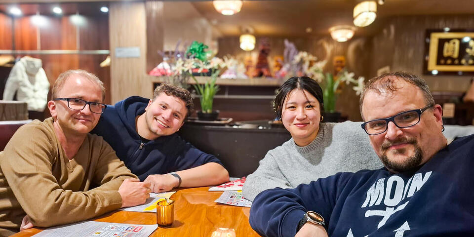 Ein junger Mann, zwei weitere Männer und eine junge Frau sitzen in einem Restaurant an einem Tisch. 
