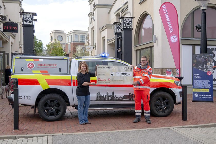 Zwei Personen halten einen Spendenscheck über 1.000 Euro vor einem Einsatzfahrzeug der Johanniter in einer Fußgängerzone.