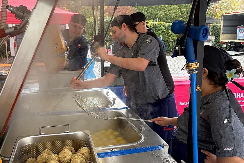  Drei Johanniter-Mitarbeitende in grauer Arbeitskleidung und schwarzen Kochmützen stehen an einer Edelstahl-Kochstation unter einem Zelt. Ein Mann hebt mit einer Schöpfkelle Knödel, während eine Frau rechts im Bild einen Sieb hält. Im Vordergrund liegt ein Metallkorb voller Knödel.