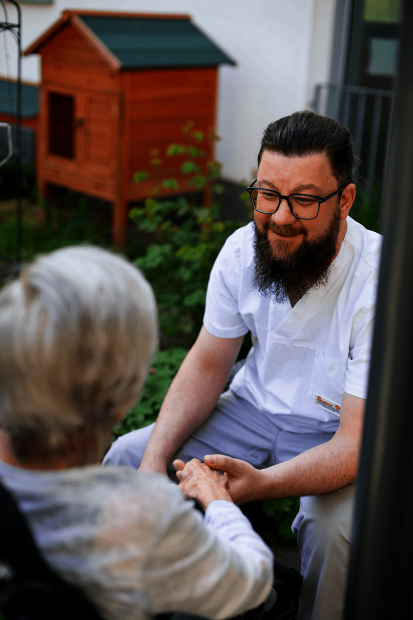 Eine männliche Pflegekraft in weißer Arbeitskleidung kniet und hält die Hände einer Person im Rollstuhl