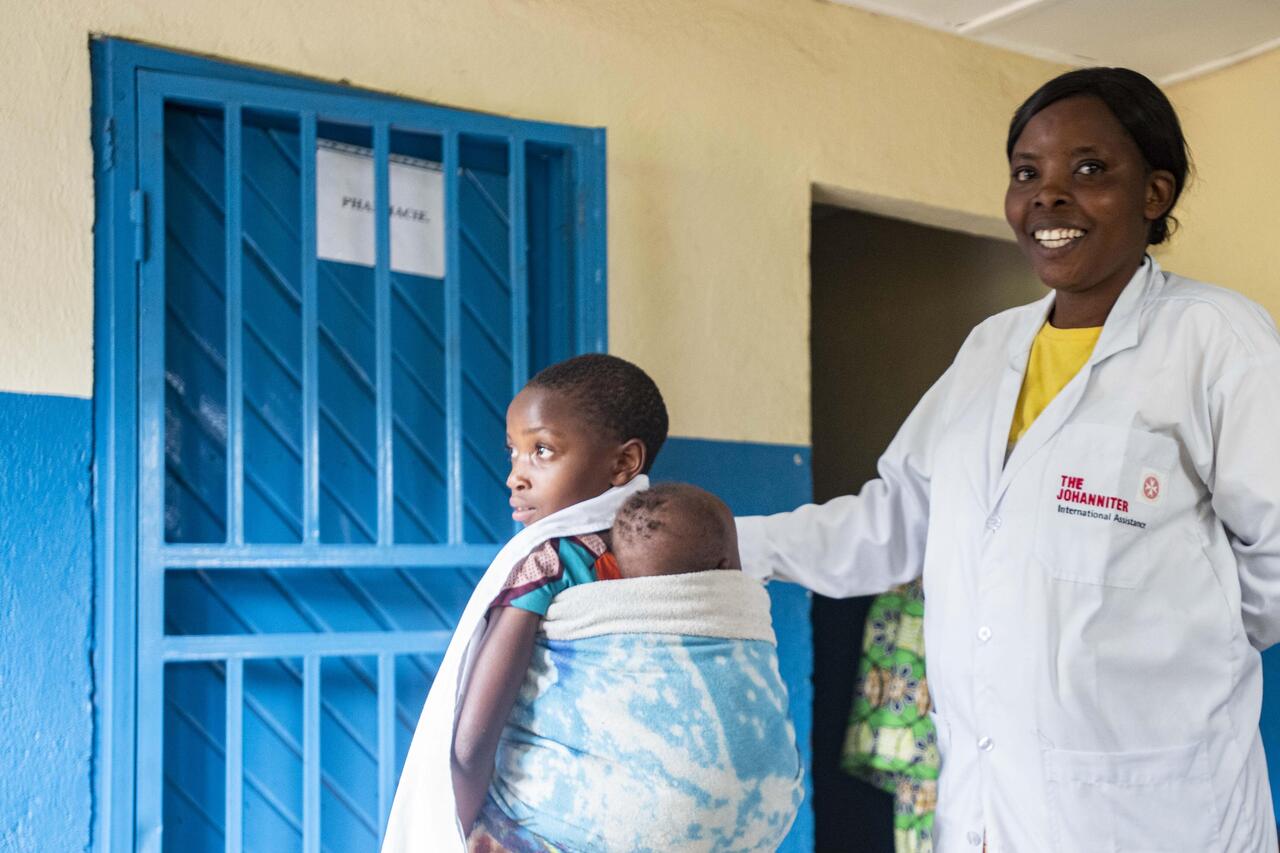 Krankenschwester in Gesundheitsstation in Nord-Kivu berührt lächelnd ein junges Kind auf dem Rücken der Mutter