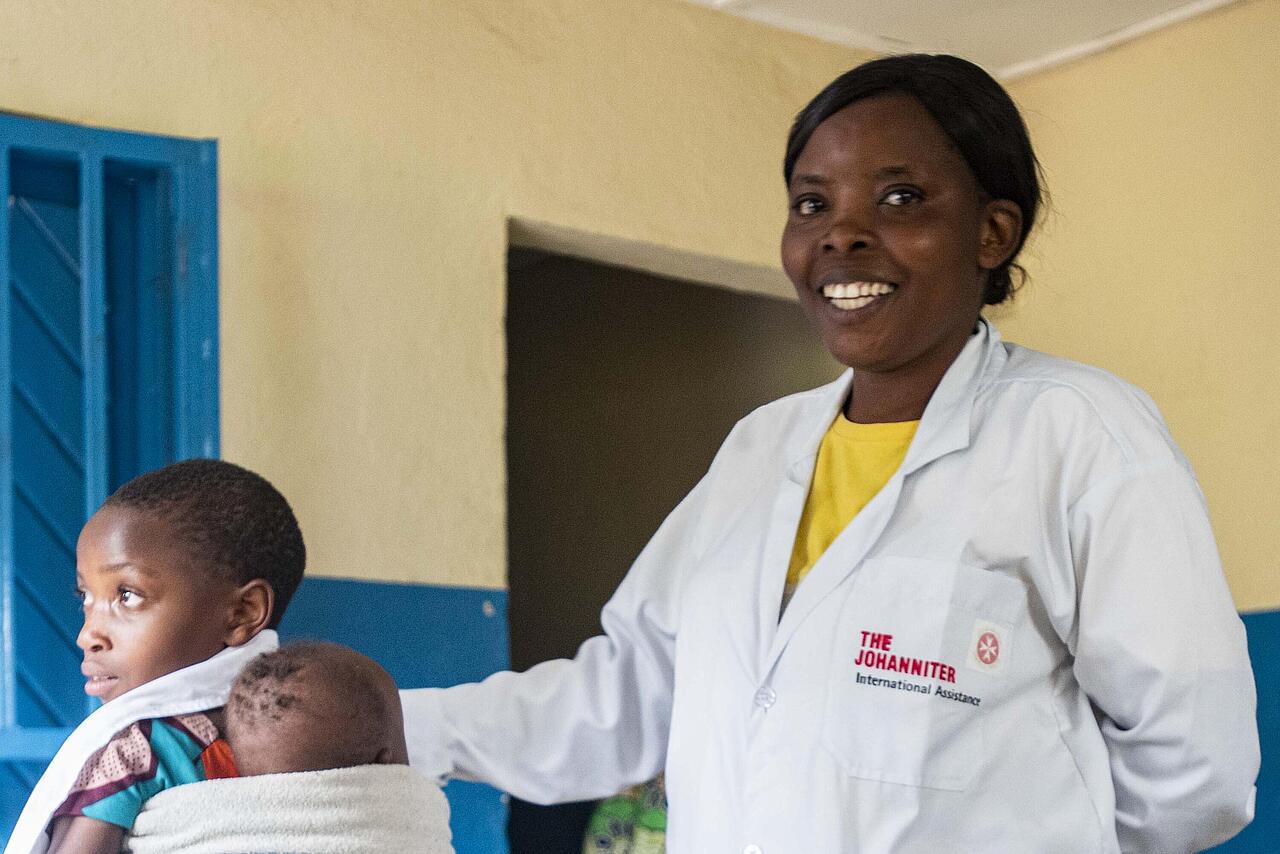 Krankenschwester in Gesundheitsstation in Nord-Kivu berührt lächelnd ein junges Kind auf dem Rücken der Mutter