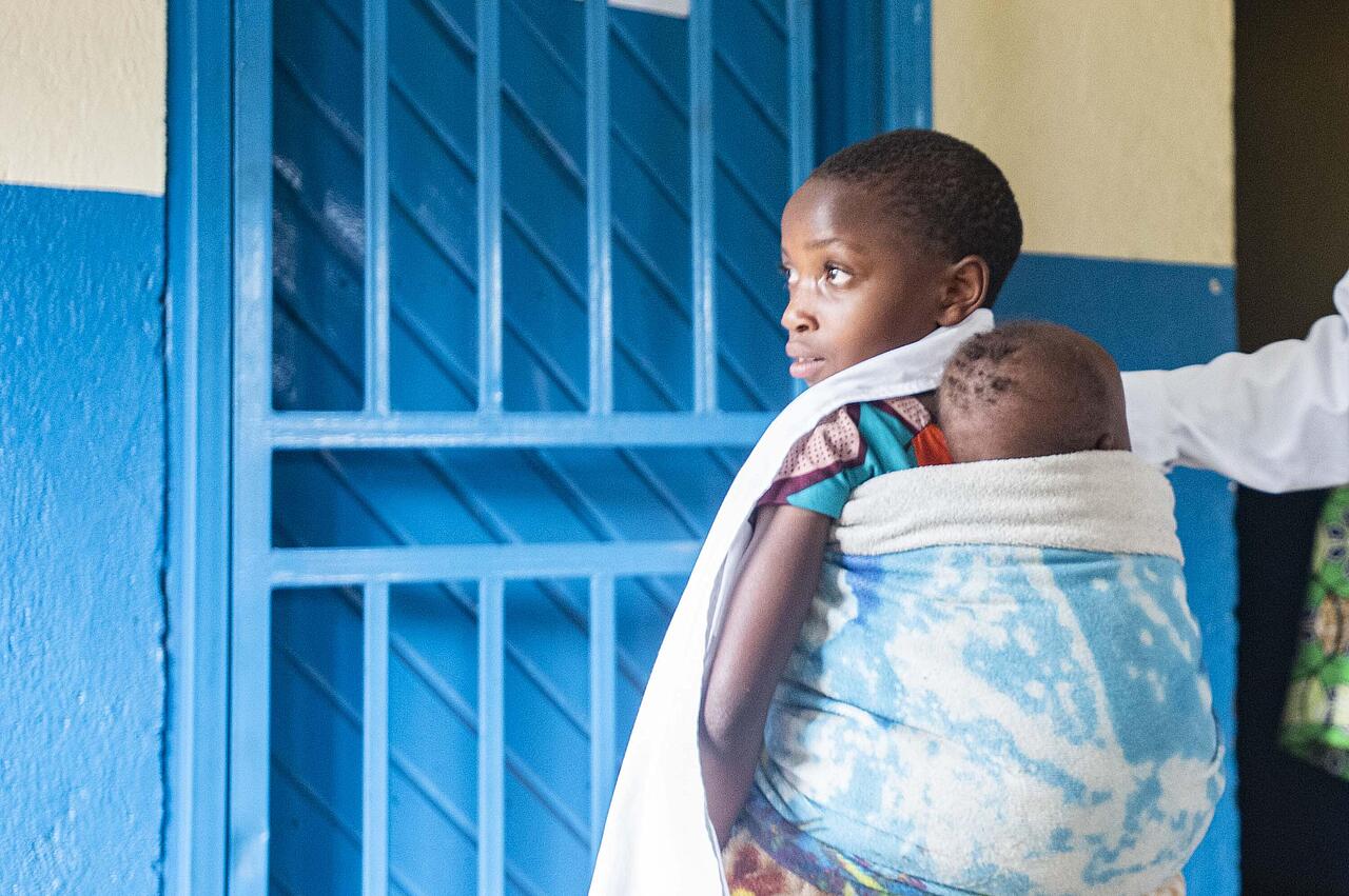 Krankenschwester in Gesundheitsstation in Nord-Kivu berührt lächelnd ein junges Kind auf dem Rücken der Mutter
