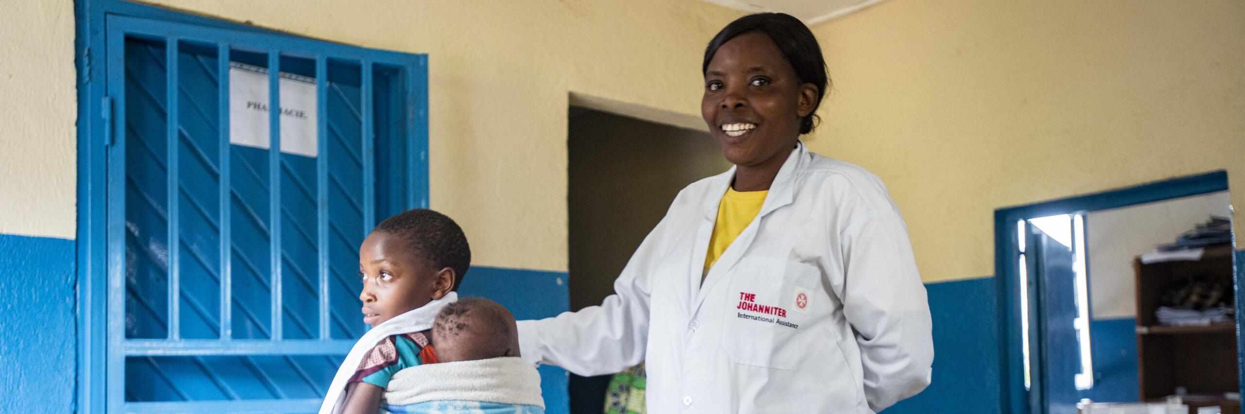 Krankenschwester in Gesundheitsstation in Nord-Kivu berührt lächelnd ein junges Kind auf dem Rücken der Mutter