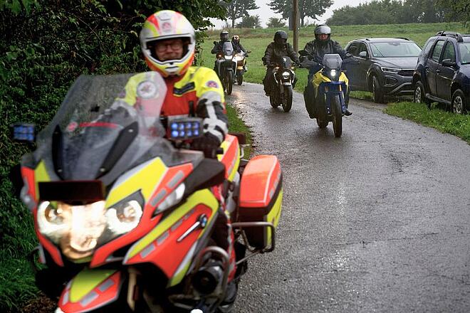 Eine Gruppe von Motorradfahrern fährt über einen nassen Asphaltweg. Im Vordergrund ein Johanniter auf Johanniter-Motorrad, im Hintergrund in dunkel gekleidete Biker auf privaten Motorrädern. 