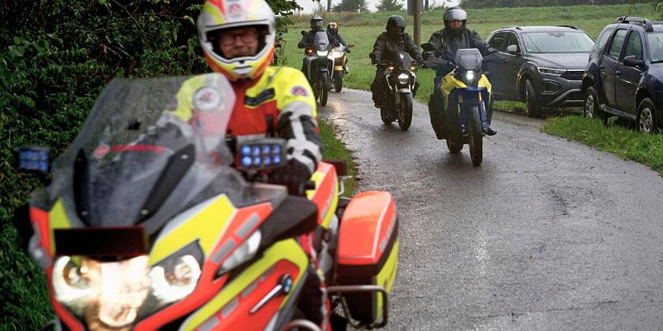 Eine Gruppe von Motorradfahrern fährt über einen nassen Asphaltweg. Im Vordergrund ein Johanniter auf Johanniter-Motorrad, im Hintergrund in dunkel gekleidete Biker auf privaten Motorrädern. 