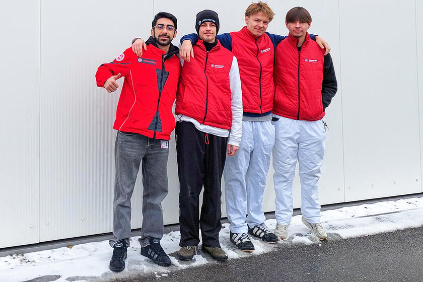 Vier junge Männer in roten Johanniter-Westen stehen Arm in Arm vor einer hellen Wand im Freien, lächeln in die Kamera, während auf dem Boden etwas Schnee liegt.