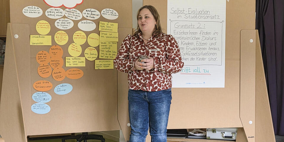 Eine Frau vor zwei großen Pinnwänden auf denen ein Plakat und Zettel kleben. Sie spricht. 