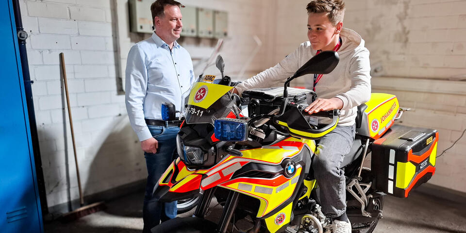 Ein Junge sitzt auf einem Motorrad in einer Halle. Ein Mann steht daneben.